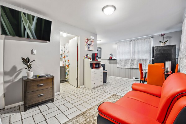 Ett modernt vardagsrum med en röd soffa, en TV som är monterad på väggen, en mikrovågsugn och en liten matplats med orange stolar.