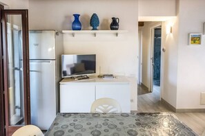 A small kitchenette with a refrigerator, a TV, and a white cabinet. There is a shelf above the cabinet with decorative items.
