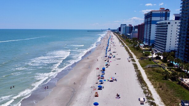 A beach with a long stretch of umbrellas and sun loungers, adjacent to high-rise buildings. - 2 Bed, 2 Bath Topsail Cottage—Walk to the beach! (Myrtle Beach)