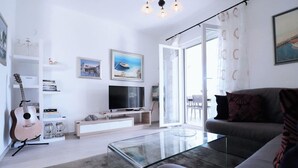 A modern living room with a glass coffee table, a grey sofa, a TV on a white stand, a guitar, and a bookshelf. - Luxury Beachfront Villa With Pool for two Families (Okrug)