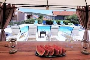 Vista da piscina com espreguiçadeiras, uma mesa com fatias de melancia e copos de vinho.