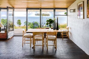 A dining area with a wooden table and chairs, a vase with flowers, and a view of the sea. - Architect designed holiday home with sea view (Gudhjem)