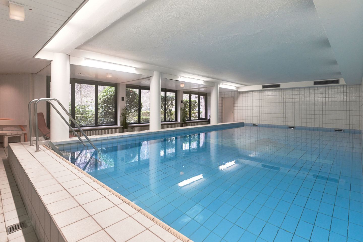 Una piscina interior con agua azul cristalina, paredes de azulejos y grandes ventanales que dejan entrar la luz natural.