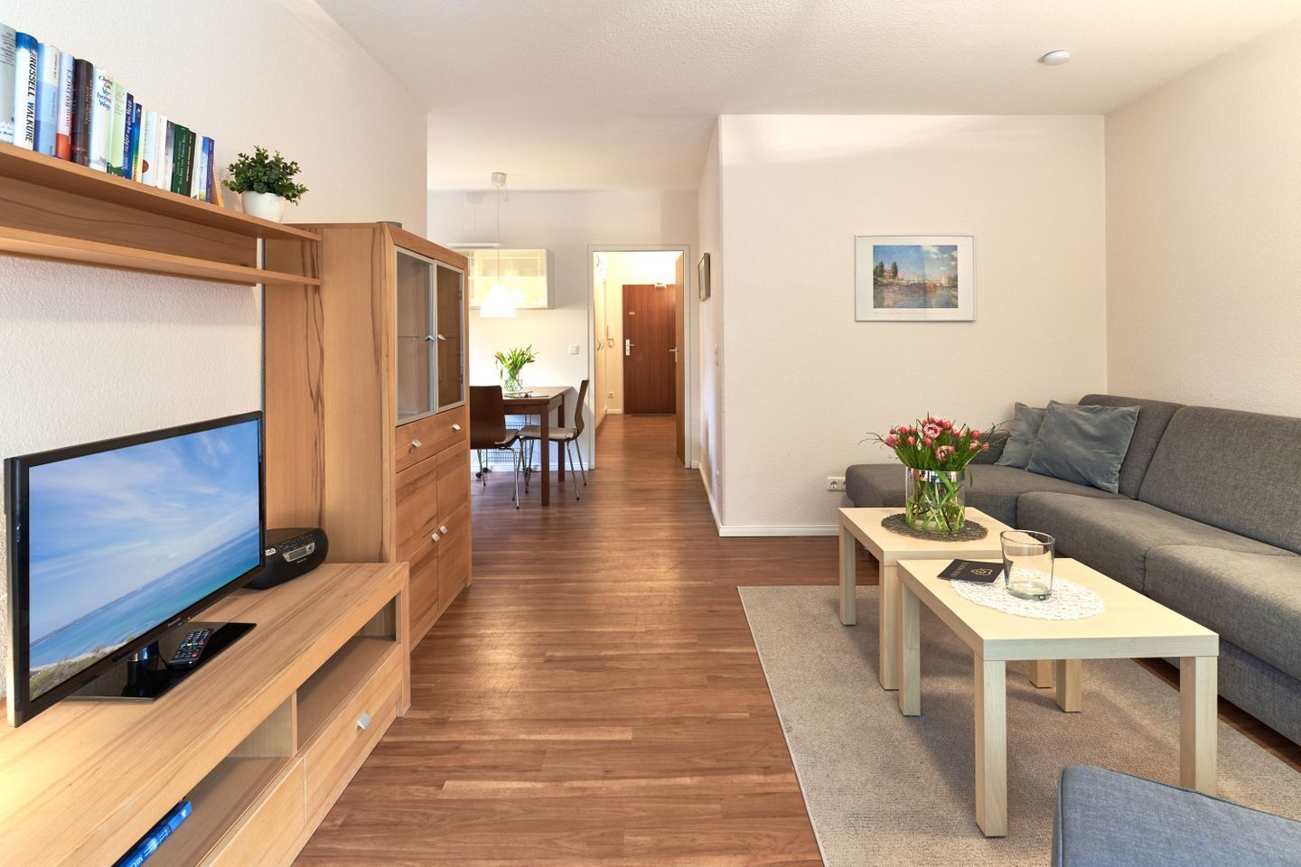 A modern living room with a flat-screen TV, wooden furniture, a grey sofa, and a dining area with a table and chairs.