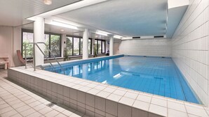 An indoor swimming pool with a tiled floor and walls, a metal pool ladder, and large windows allowing natural light.