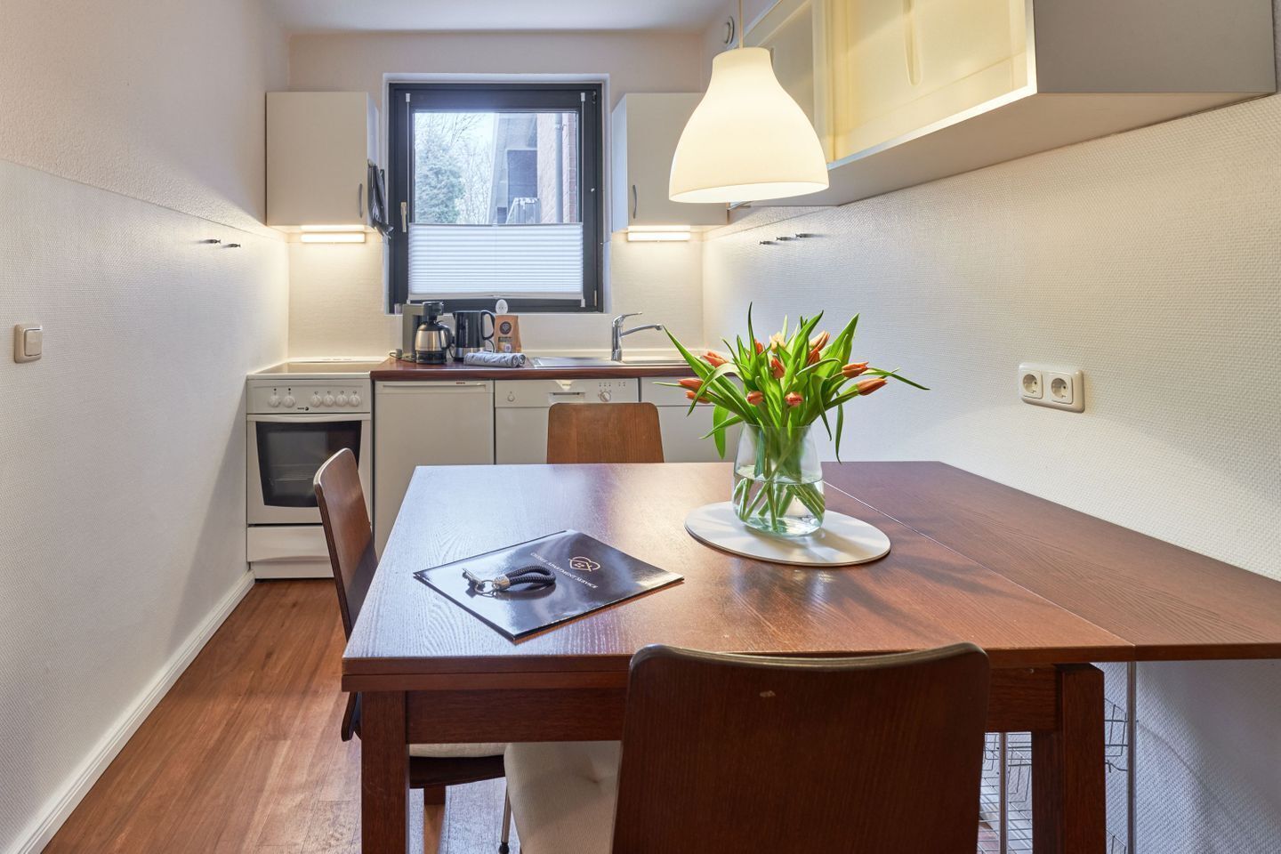 Una cocina compacta con comedor, una ventana con persianas y un florero con flores sobre la mesa.