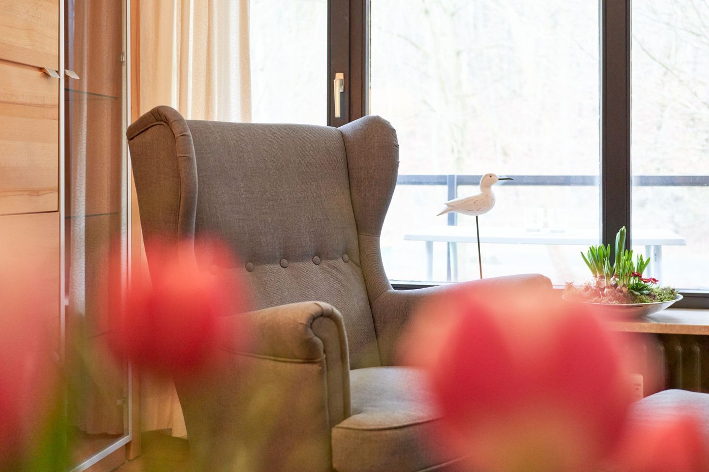 A room with a grey upholstered armchair, a window with a view of trees, and a small table with a plant.