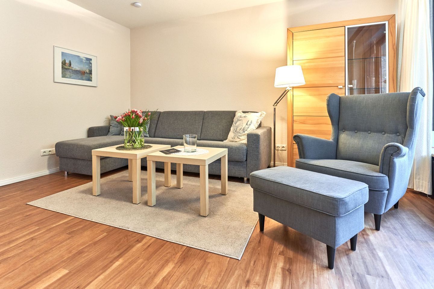 Een moderne woonkamer met een grijze bank, twee houten bijzettafeltjes, een grijze fauteuil en een vloerlamp.