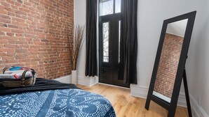 Una habitación con pared de ladrillo, cama con colcha estampada, caballete de marco negro y puerta con cortinas negras.