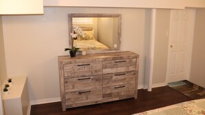 A bedroom with a wooden dresser, a mirror, a bed with floral bedding, and a white door. - Spacious 3 Bedroom walkout Basement Apartment (Ajax)