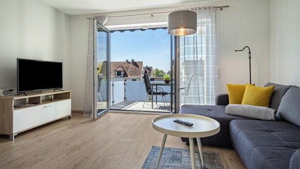 Un salon moderne doté d’un téléviseur à écran plat, d’un canapé avec des coussins, d’une petite table ronde et d’un balcon offrant une vue sur les bâtiments.