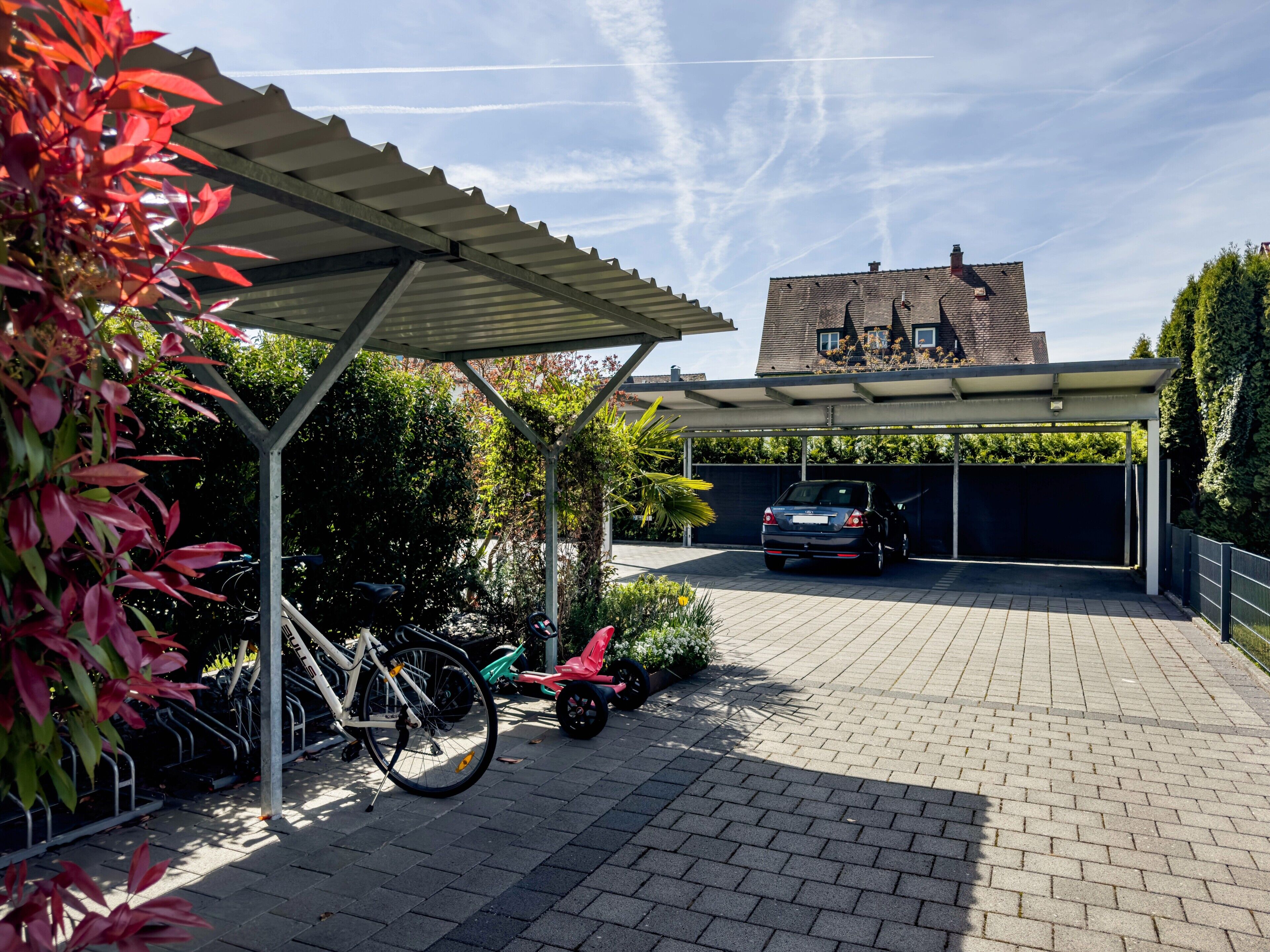 A covered parking area with a car and bicycles, a brick pathway, and a house in the background.
