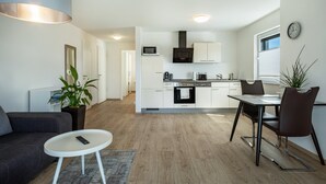 Une cuisine moderne avec des meubles blancs, un four à micro-ondes et une cuisinière. On y trouve un coin repas avec une table et des chaises, un canapé gris et une petite table d’appoint blanche.