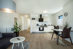 Una cocina moderna con armarios blancos, microondas y estufa. Hay un comedor con una mesa y sillas, un sofá gris y una pequeña mesita auxiliar blanca.