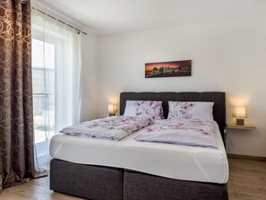En snyggt bäddad säng med blommigt sänglinne, en målning med ett stadsmotiv ovanför och ett fönster med gardiner.