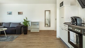 Una sala de estar moderna con un sofá, una mesa de centro, una planta, una estantería, una cocina con microondas, cafetera y horno.