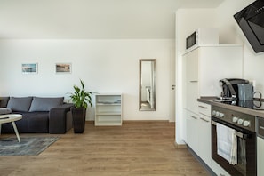 Un salon moderne comprenant un canapé, une table basse, une plante, une bibliothèque, une cuisine équipée d’un four à micro-ondes, d’une machine à café et d’un four.