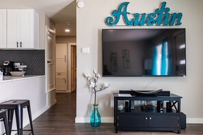 Cocina moderna con gabinetes blancos, encimera negra y salpicadero. Un televisor grande está montado en la pared sobre un mueble de entretenimiento negro.