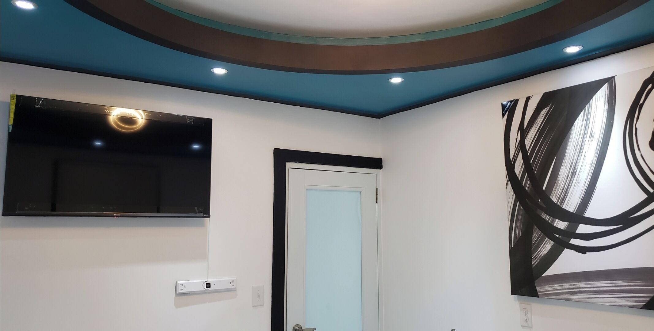 A modern room with a flat-screen TV mounted on the wall, a door with a light blue panel, and a large abstract painting on the wall.
