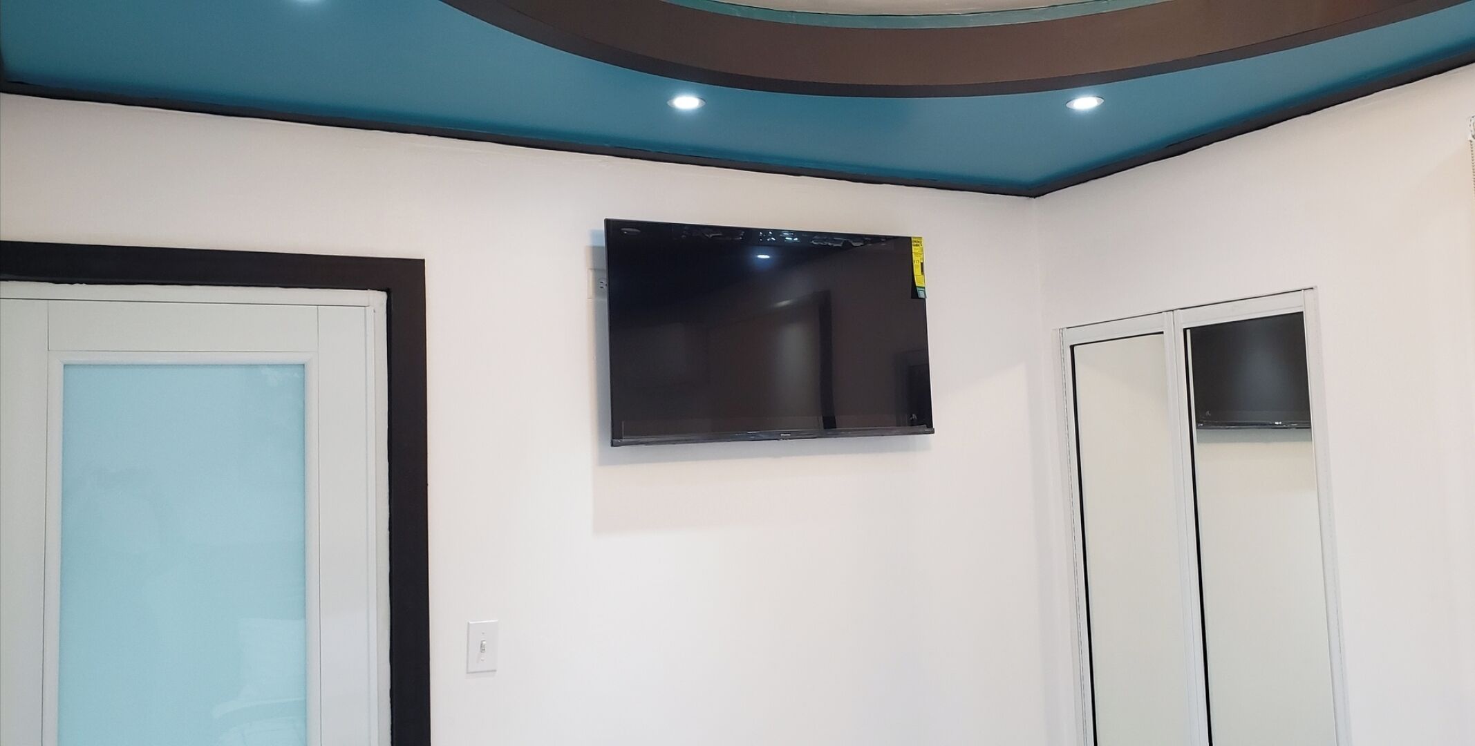 A room with a flat-screen TV mounted on the wall, a door with a light blue panel, and a white wall.
