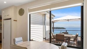 A dining area with a round table and four chairs, a clock on the wall, and a view of the sea through large windows. - The Alonnah' Beach House - Main House & Unit (Alonnah)