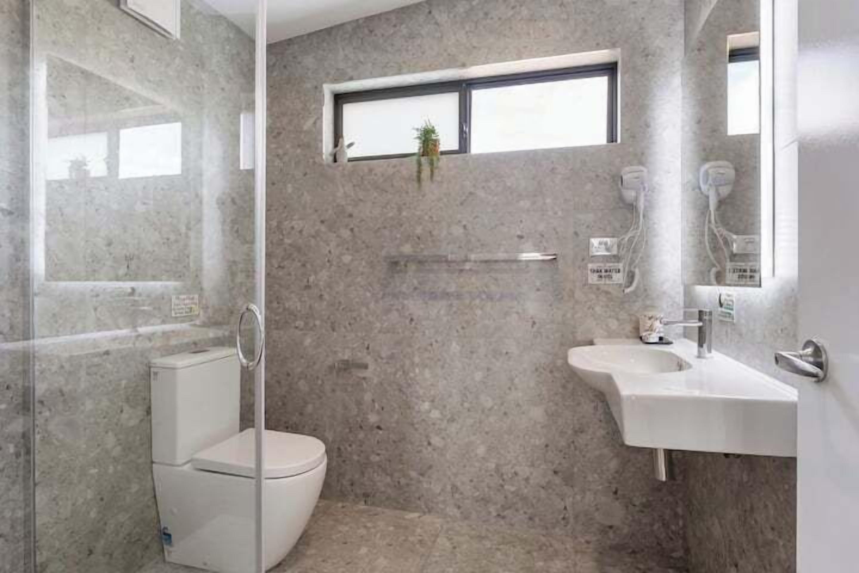 Et badeværelse med bruseniche i glas, toilet, vask og et lille vindue med en plante.