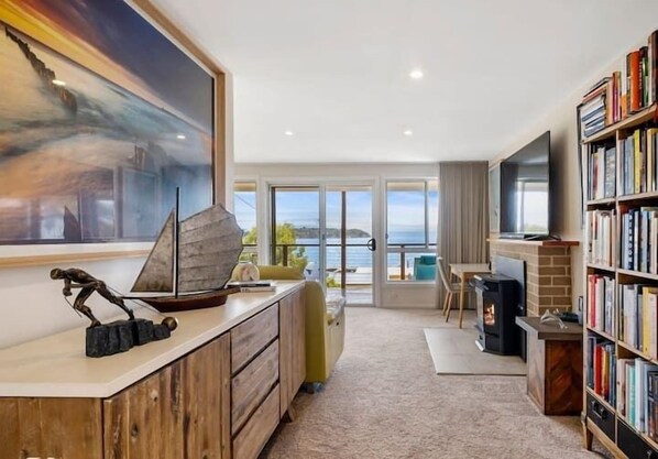 A modern living room with a large painting, a fireplace, a bookshelf, and a view of the sea through large windows. - The Alonnah' Beach House - Main House & Unit (Alonnah)