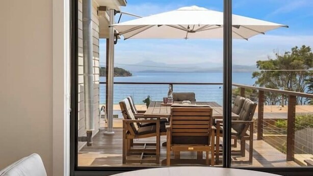 A balcony with a wooden table and chairs, a white umbrella, and a view of the sea. - The Alonnah' Beach House - Main House & Unit (Alonnah)