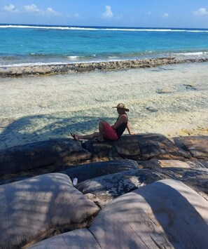 En person sidder på en sten ved en klar strand med udsigt over havet.