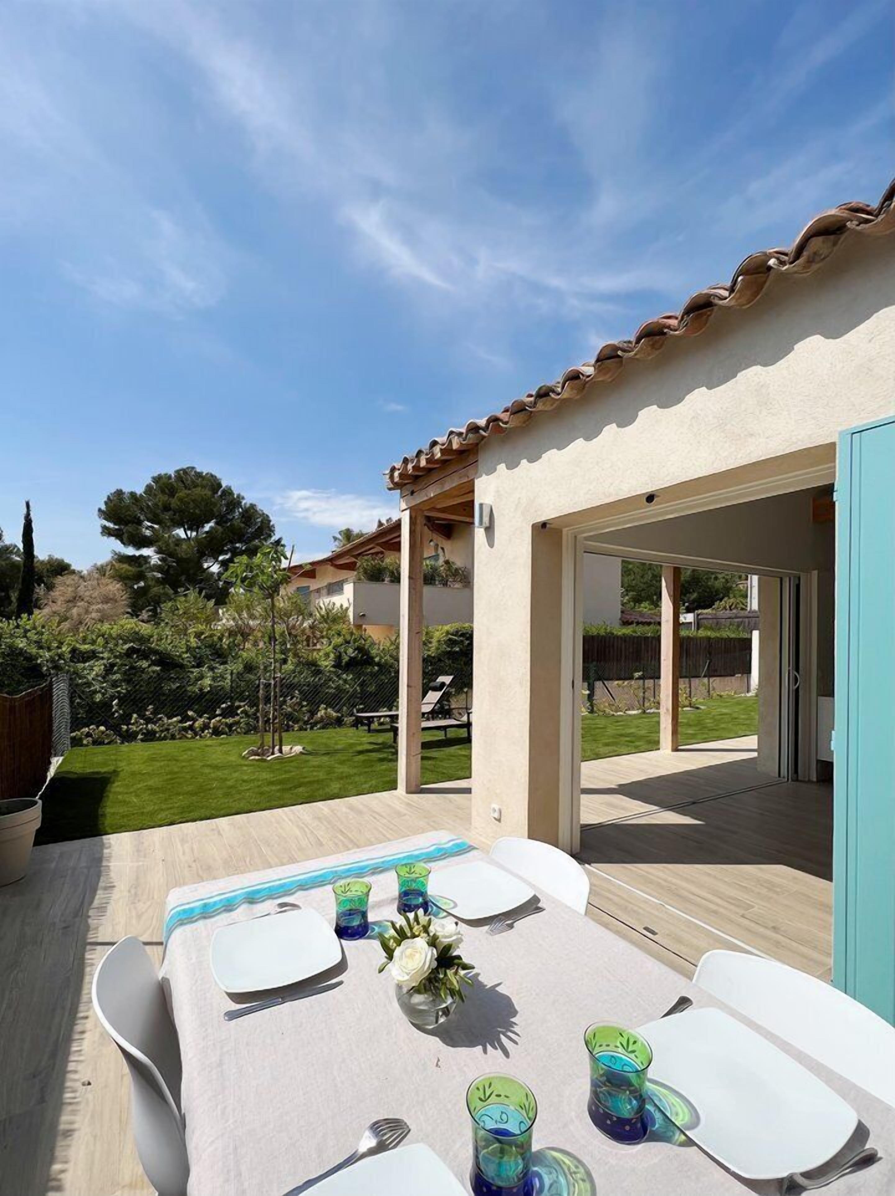 An outdoor patio with a table set for four, surrounded by a garden and a building with a tiled roof.