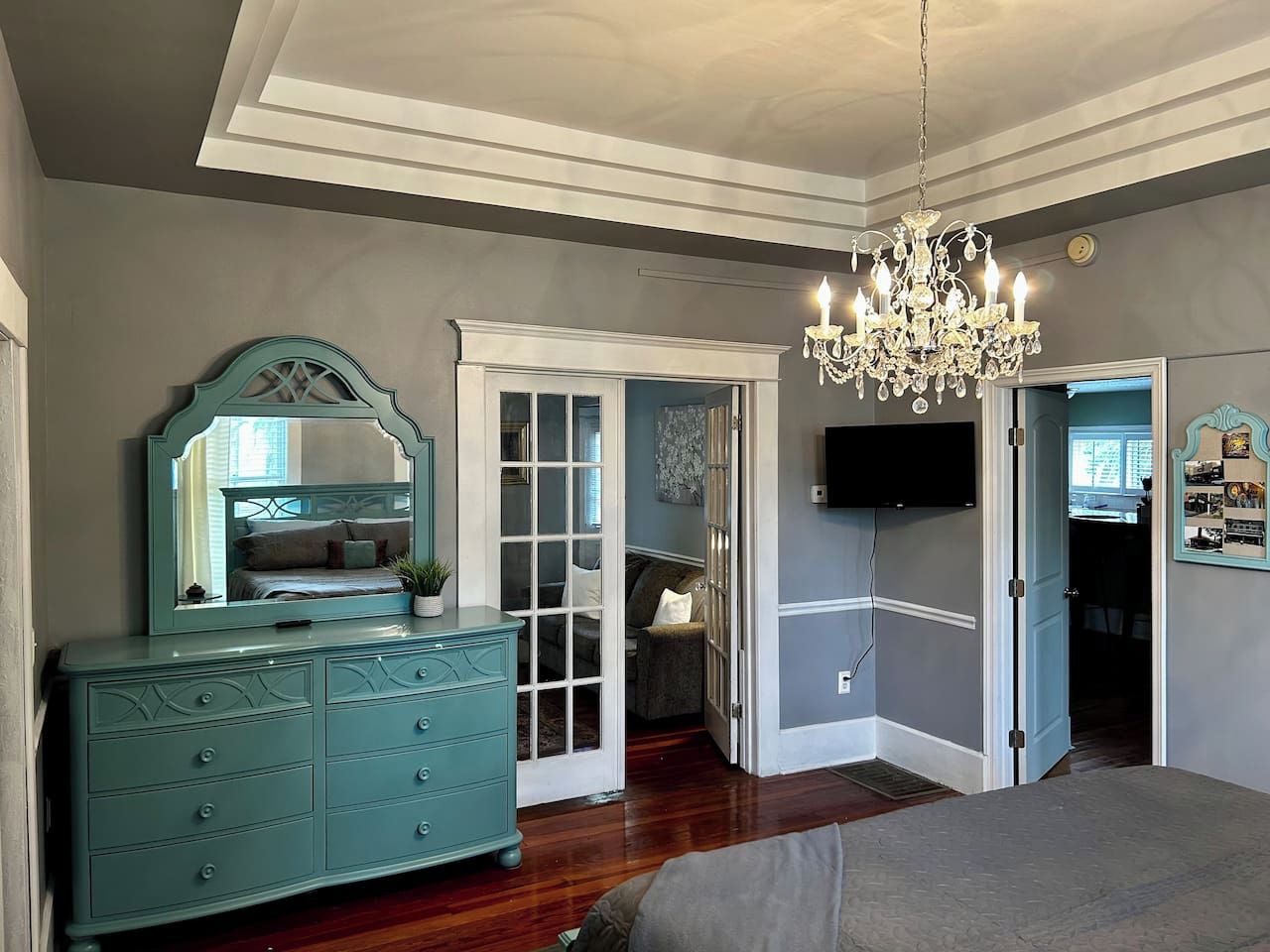 A bedroom with a large bed, a dresser with drawers, a mirror, a chandelier, and a television mounted on the wall.