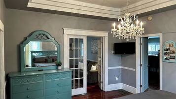 A bedroom with a large bed, a dresser with drawers, a mirror, a chandelier, and a television mounted on the wall.