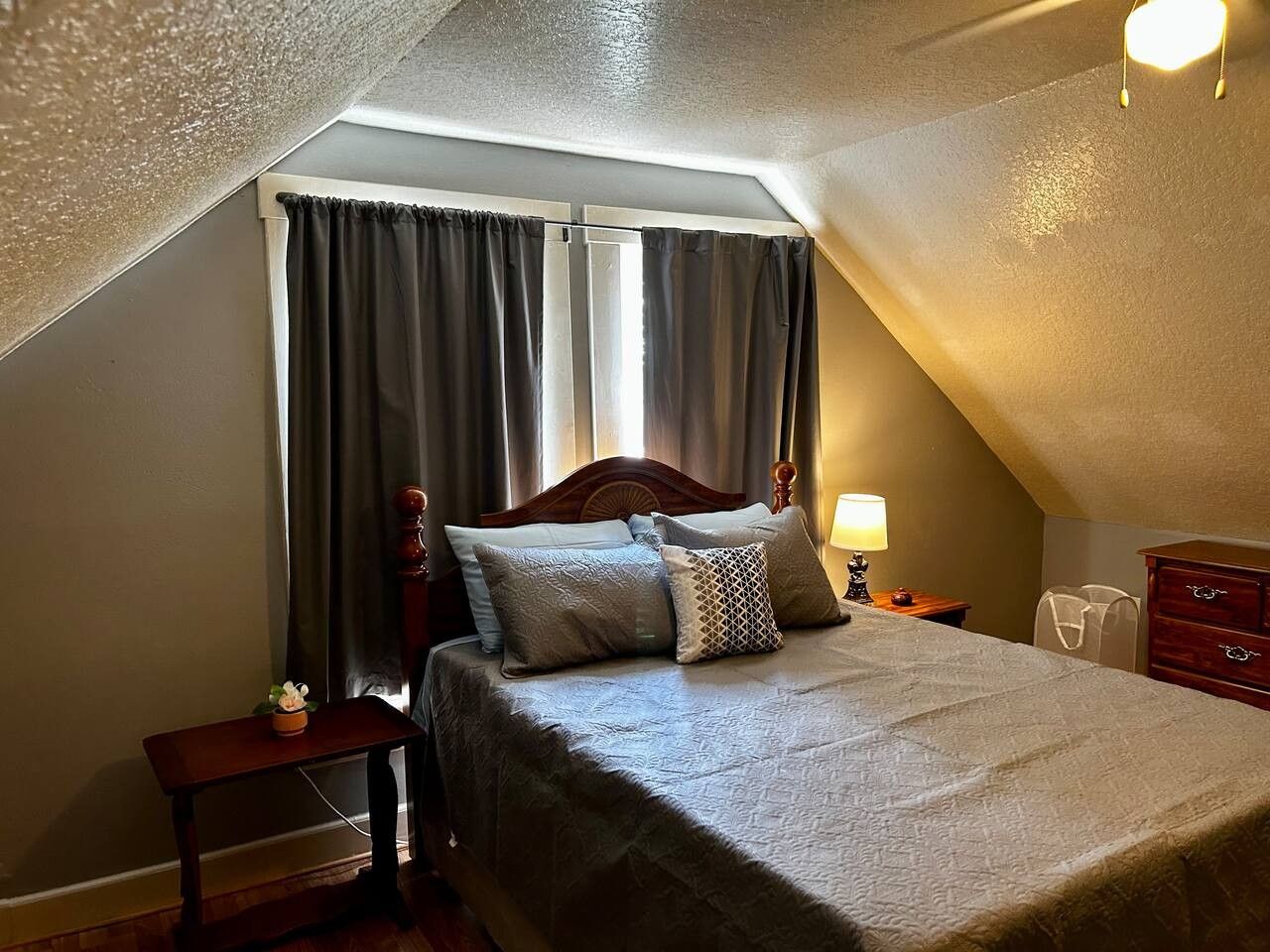 A bedroom with a sloped ceiling, a bed with a wooden headboard, a bedside table with a lamp, and a dresser.