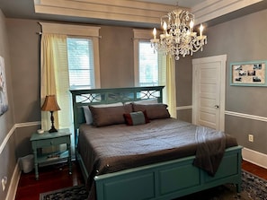 A bedroom with a bed, a nightstand, a lamp, a chandelier, and a door.