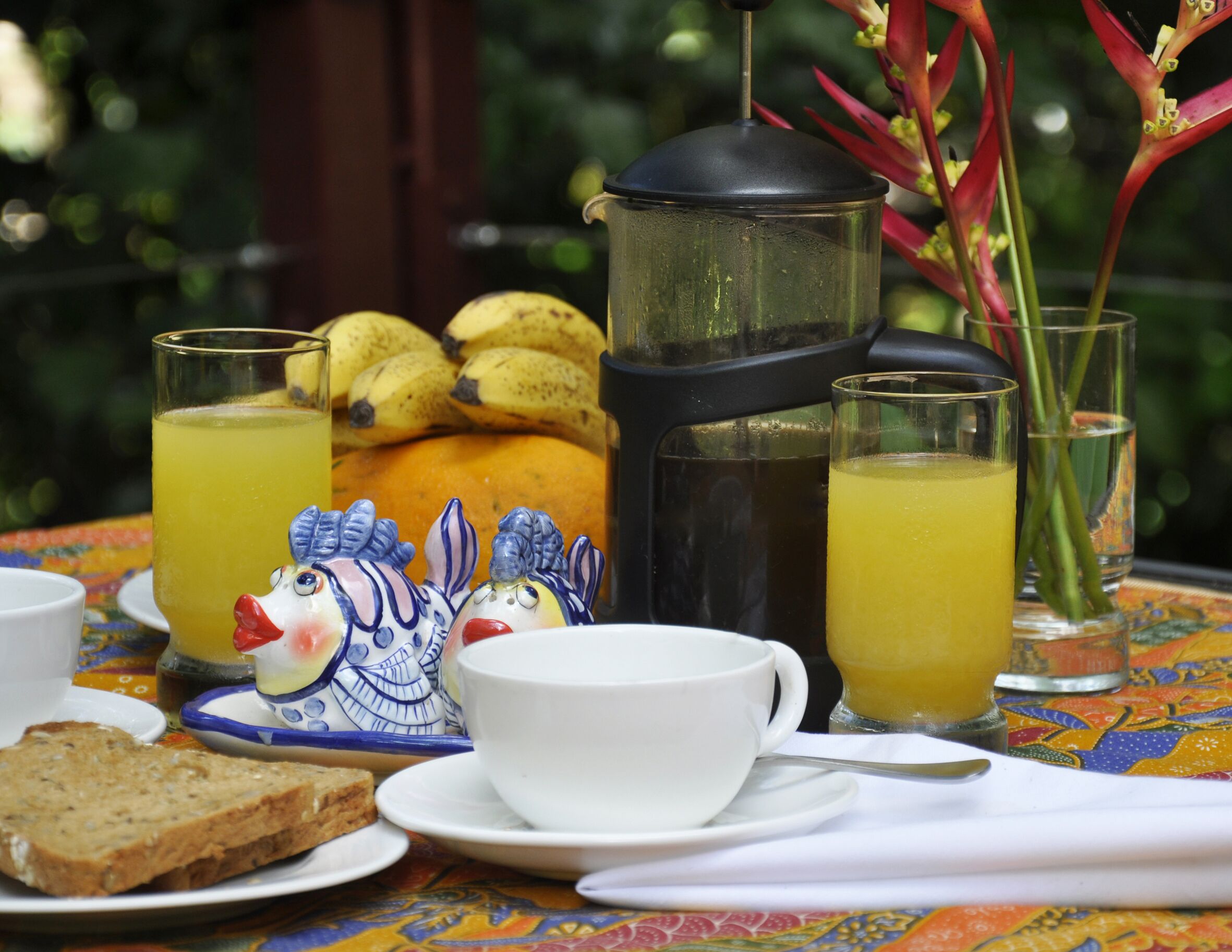 Un desayuno con una cafetera, dos vasos de zumo de naranja, un plato de plátanos, un pato de cerámica, una taza y una rebanada de pan.