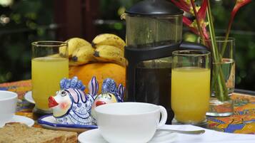 Un desayuno con una cafetera, dos vasos de zumo de naranja, un plato de plátanos, un pato de cerámica, una taza y una rebanada de pan.