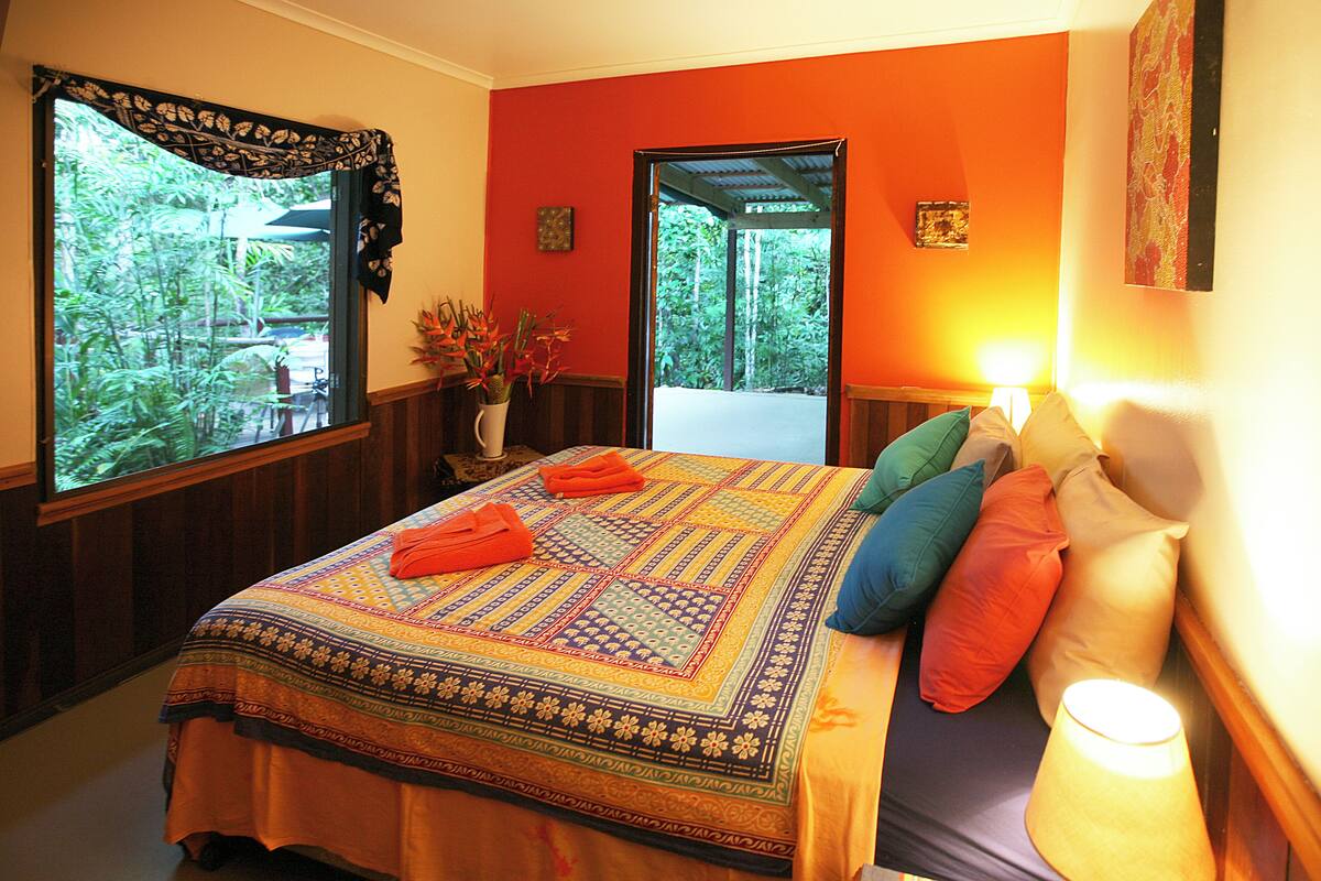 A bedroom with a large bed, colorful pillows, a nightstand with a lamp, and a window with a view of greenery.