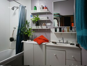 Un baño con bañera, lavabo, espejo, repisas con artículos de aseo y una planta en maceta.