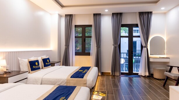 A hotel room with two beds, a desk, a chair, a small table, a mirror, and a balcony with a view of the outside. - SUNRISE Hotel Bac Lieu (Bac Lieu)