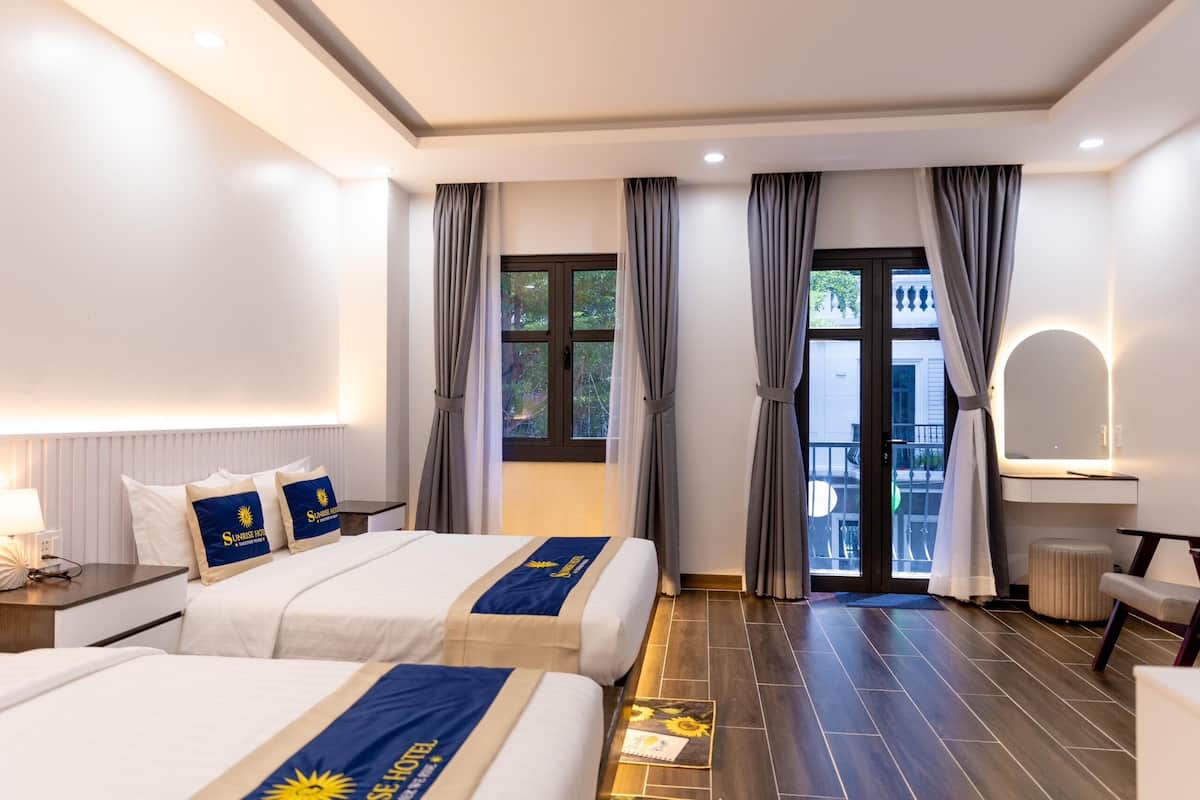 Quarto de hotel com duas camas, uma escrivaninha, uma cadeira, uma mesinha, um espelho e uma varanda com vista para o exterior.