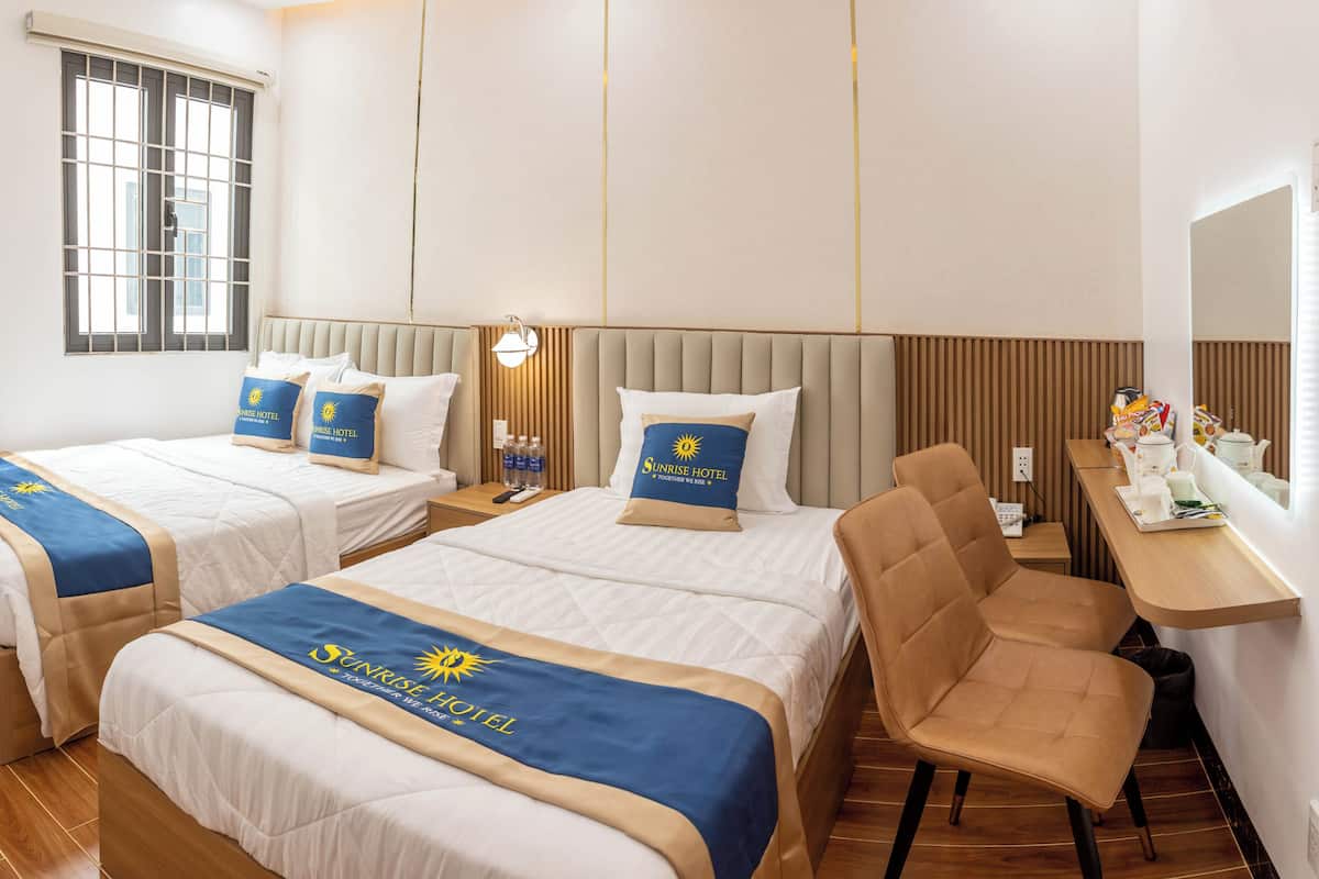 Quarto de hotel com duas camas, cada uma com manta azul e bege com o logotipo do Sunrise Hotel. Apresenta uma mesa de cabeceira de madeira com abajur, uma cadeira e uma pequena mesa com bule e xícaras.