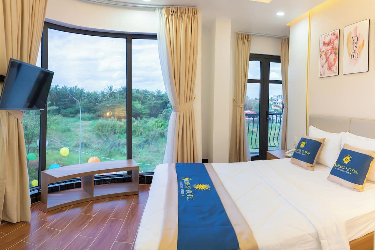 Quarto de hotel com uma cama grande, uma escrivaninha com cadeira, televisão e vista para o verde através de janelas amplas.