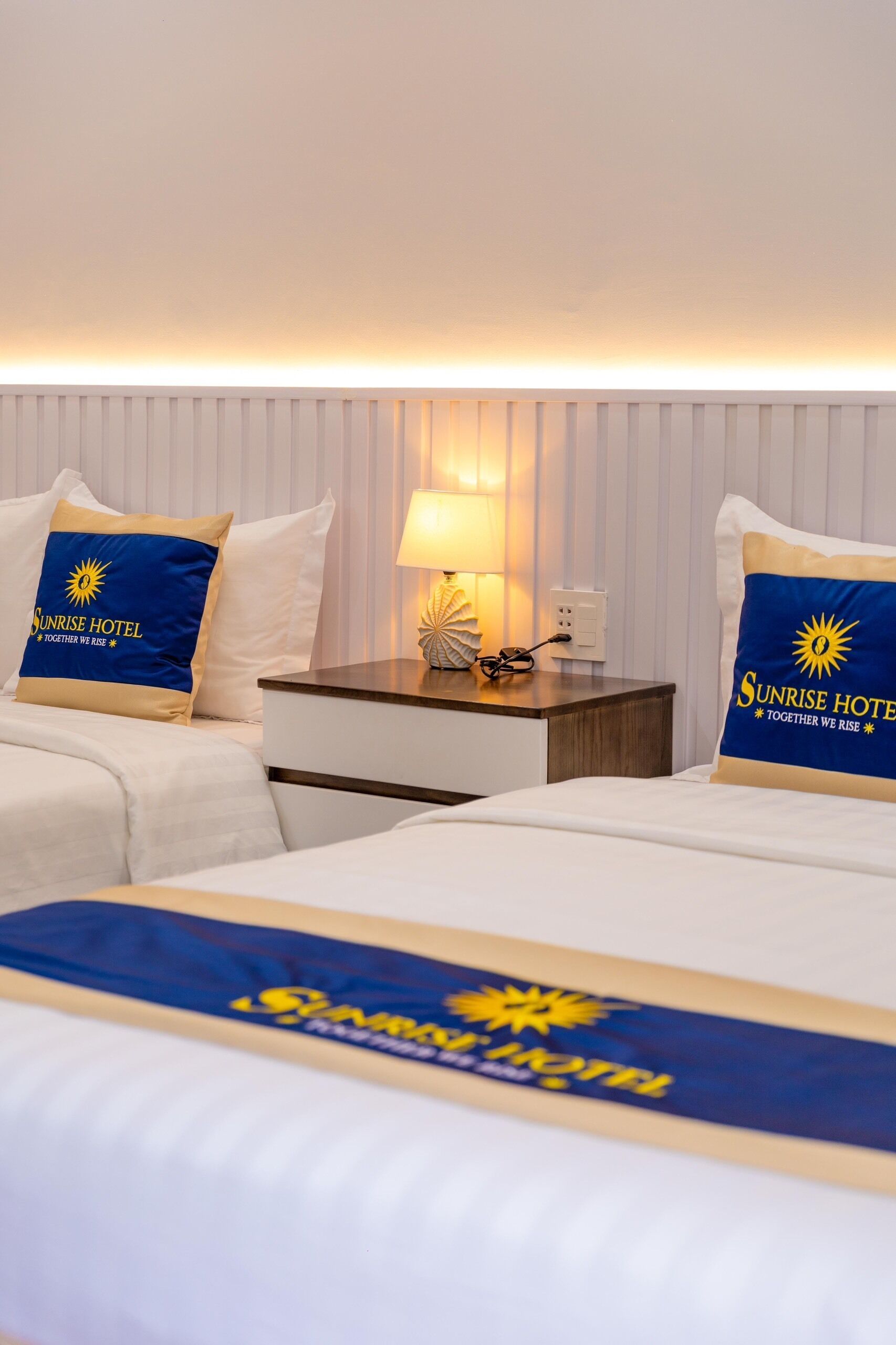 A hotel room with two beds, each with a Sunrise Hotel pillow, a bedside table with a lamp, and a wall-mounted light.