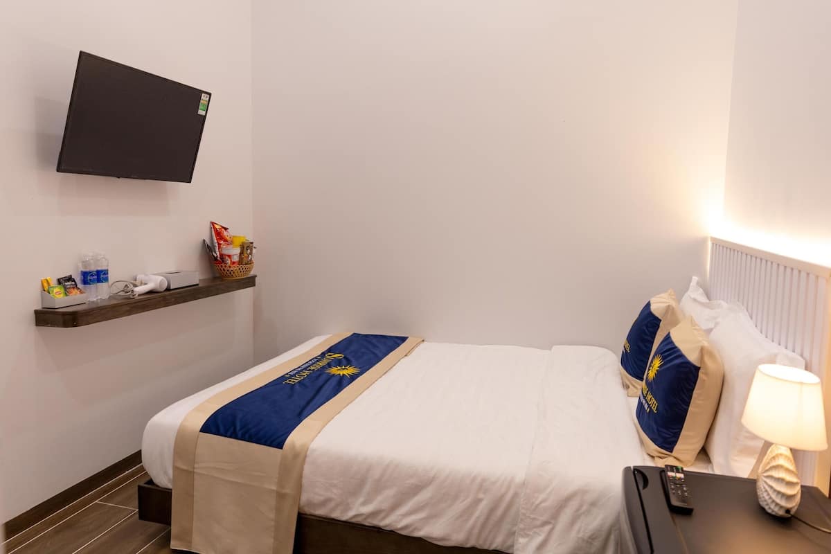 Quarto de hotel com cama, televisão de tela plana na parede, mesa de cabeceira com luminária e prateleira com artigos de higiene pessoal.