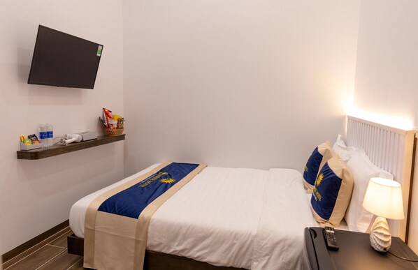 A hotel room with a bed, a flat-screen TV mounted on the wall, a bedside table with a lamp, and a shelf with toiletries.