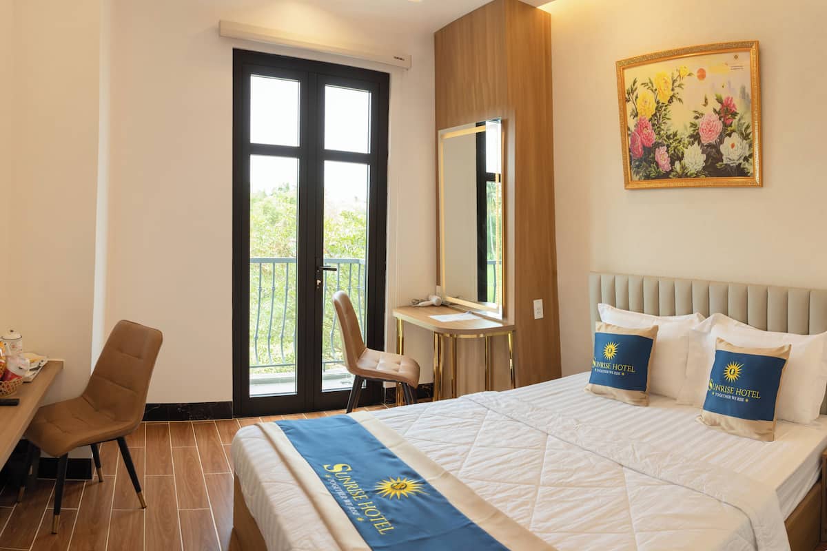 Quarto de hotel com cama, escrivaninha, cadeira, uma janela grande e um quadro na parede.