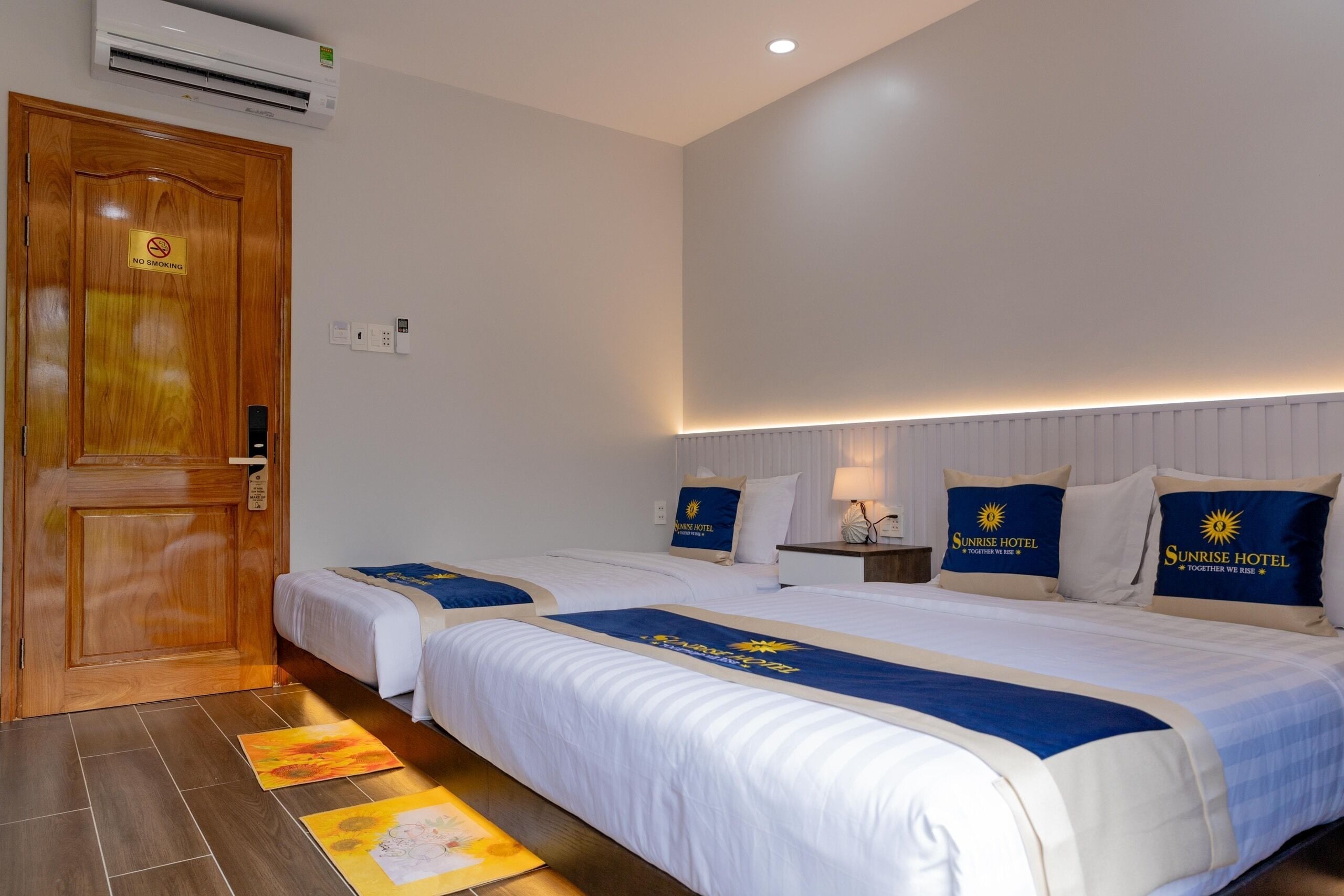 A hotel room with two beds, a wooden door, and a "No Smoking" sign.