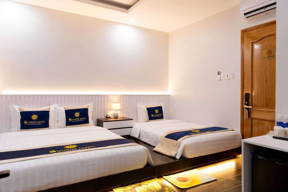 Quarto de hotel com duas camas, cada uma com almofada do logotipo Sunrise Hotel, uma mesa de cabeceira com abajur, uma porta de madeira e uma pequena escrivaninha com controle remoto e garrafas de água.