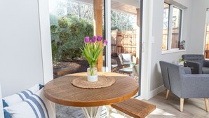 Un tavolo rotondo in legno con un sottopiattino intrecciato, un vaso con fiori viola e vista su un giardino esterno attraverso ampie vetrate.