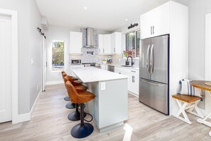 A modern kitchen with white cabinetry, a stainless steel refrigerator, and a central island with bar stools. - Beautiful new home, short stroll to the ocean. (Sooke)